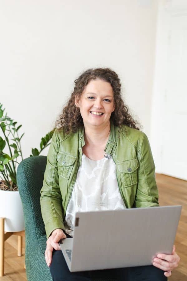 Claudia sitzt auf einem Sessel, hält einen Laptop in der Hand und lächelt